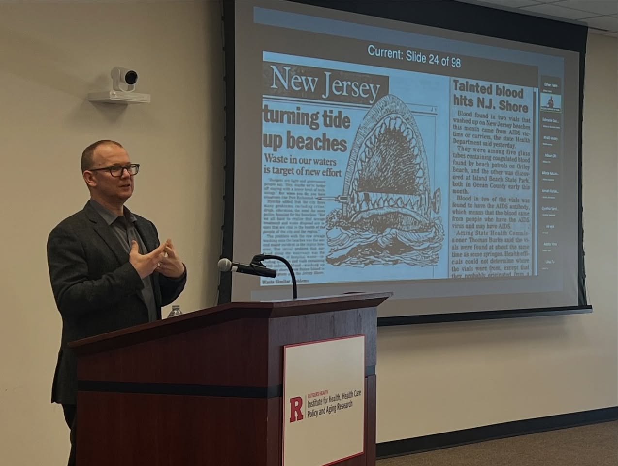 Jeremy Greene stands at a lecture in a suit with a slide on the projector showing a cartoon shark eating a syringe from a New Jersey press clipping.