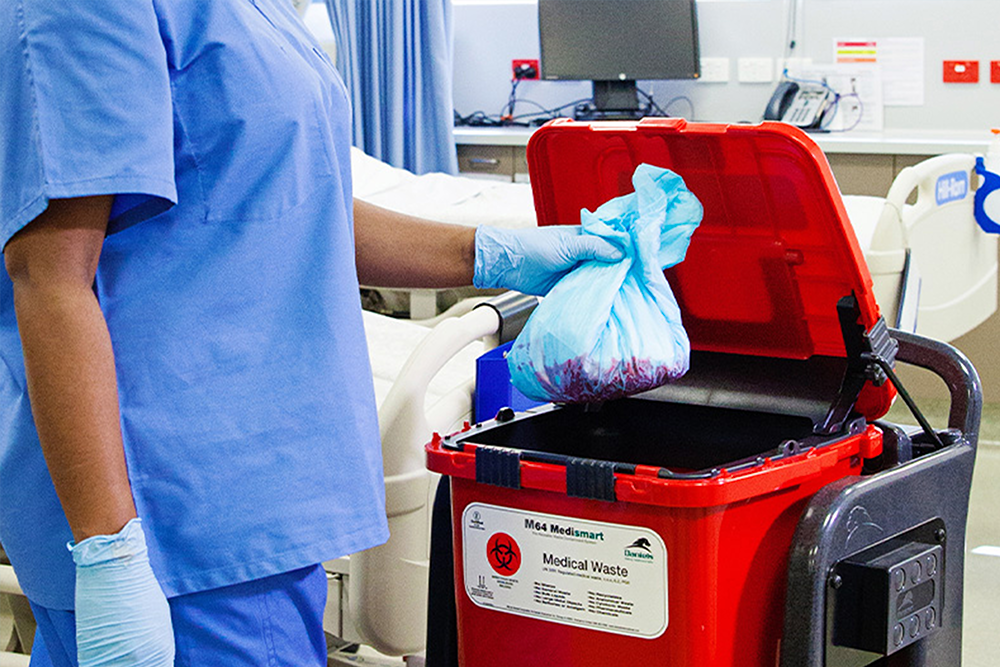 A hospital worker in blue scrubs puts a plastic bag of medical waste into a red bin.