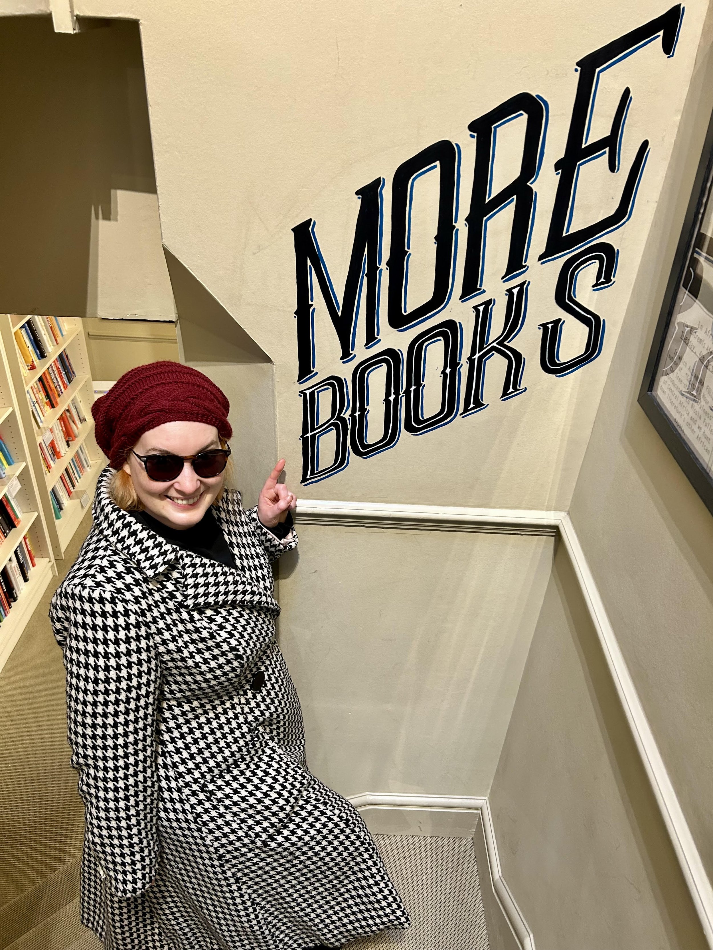 Miranda Melcher stands on a staircase pointing at the words "More Books" which have been printed on the wall by her side.
