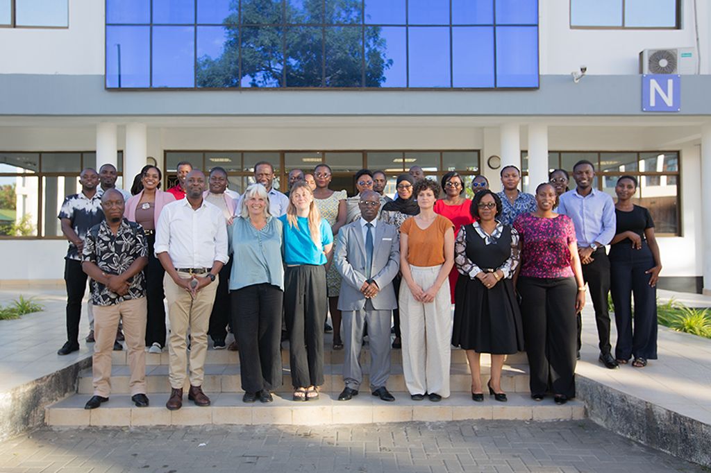 A group of healthcare professionals and academics stand in front of MUHAS in the sunshine.