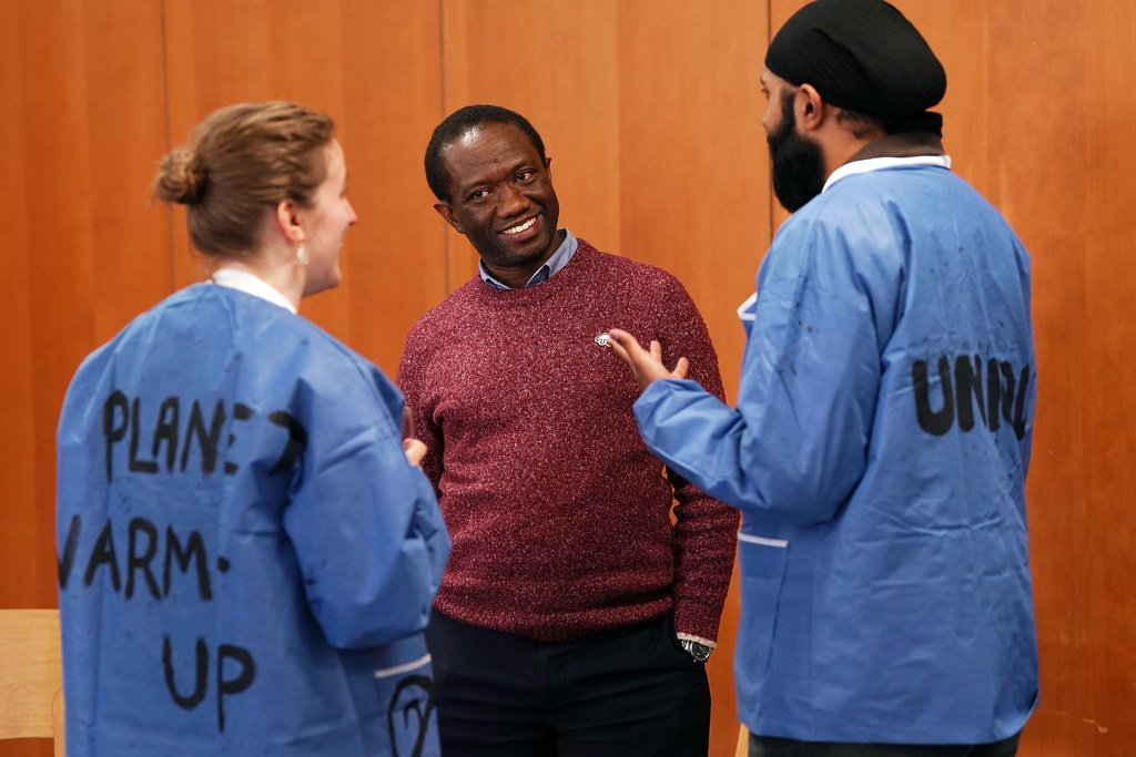 Two students wearing disposable blue jackets decorated with the words "Planet Warm-Up" and "Unique" talking to Amani Anaeli 