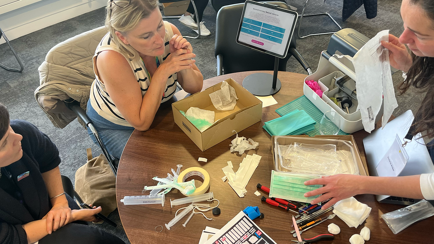 Three workshop participants are sorting through various components of a medical device with a tablet showing a series of data tables in front of them. 