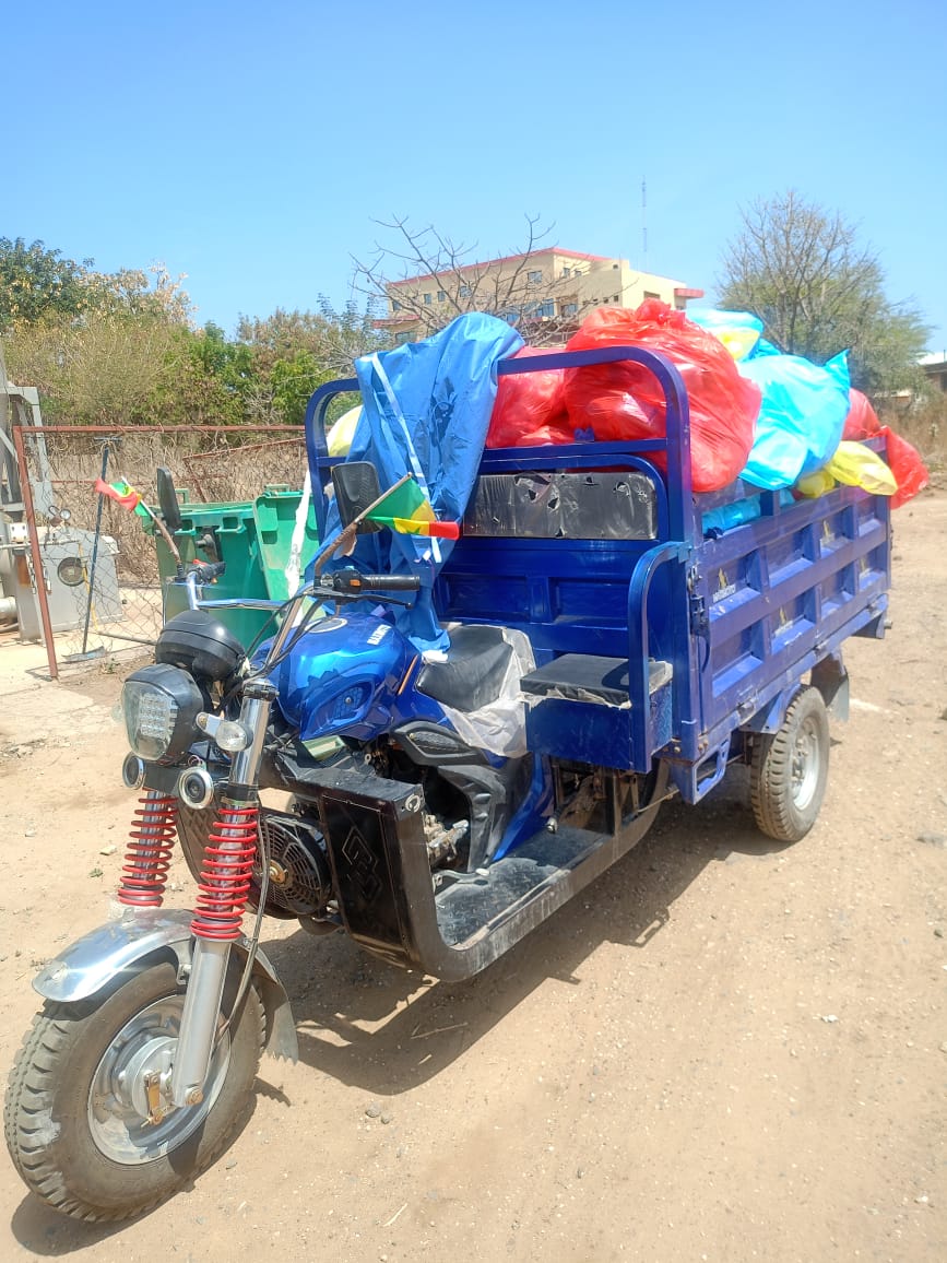 A motorbike trike is fitted with a blue trailer that filled with plastic bags of waste. 