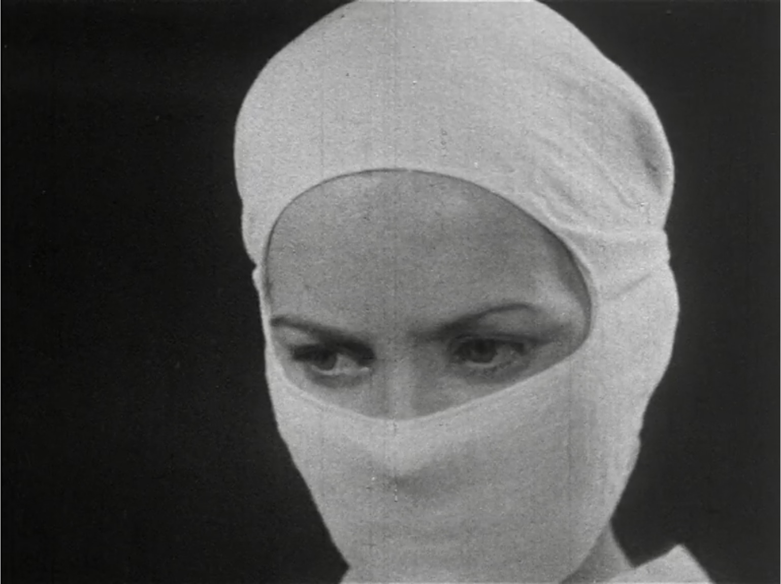 A black and white still of a nurse in a white face mask, only her eyes are visible. 