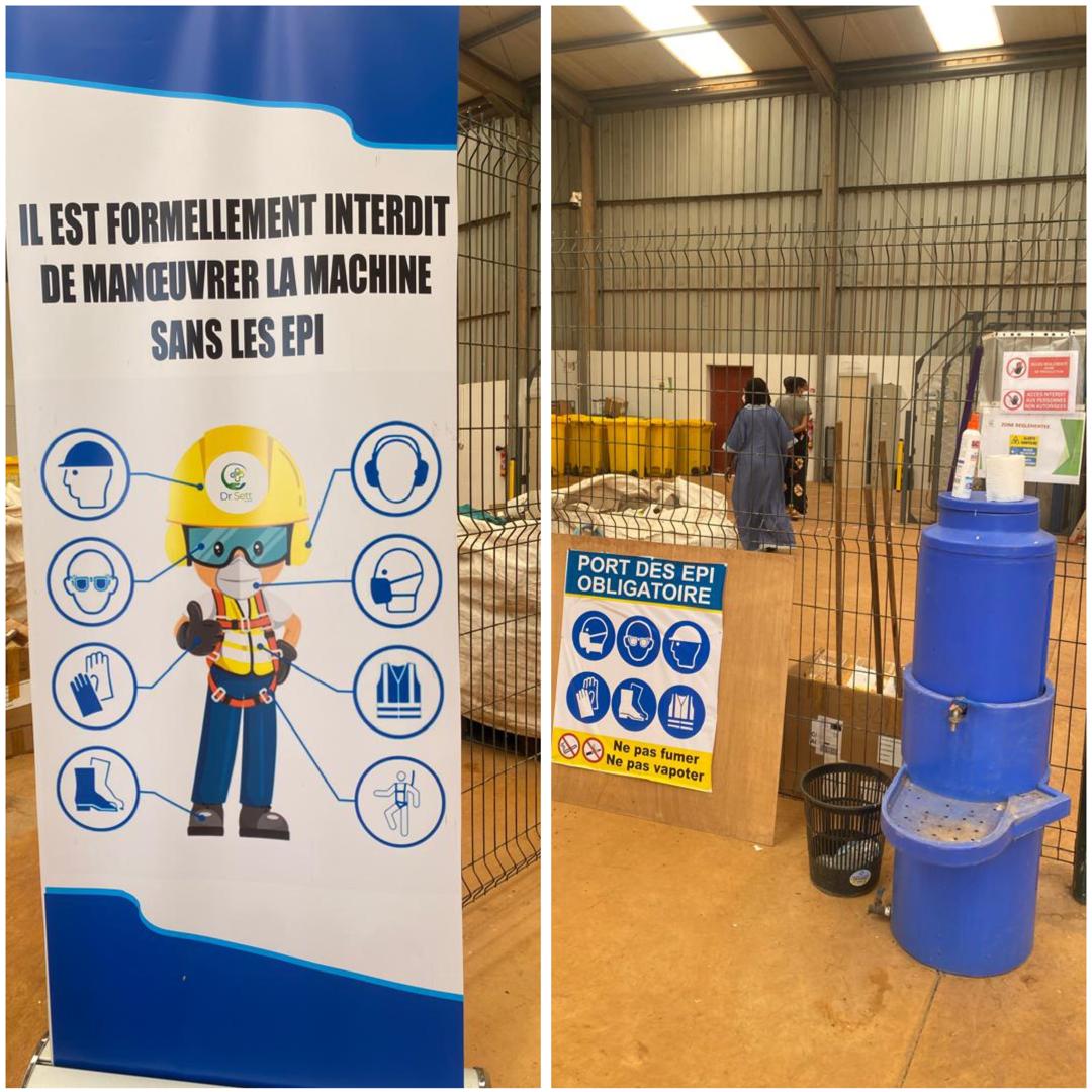 A large poster illustrating PPE needed to enter the factory. A metal wire fence separates the room with a blue plastic water hand washing station in the foreground.
