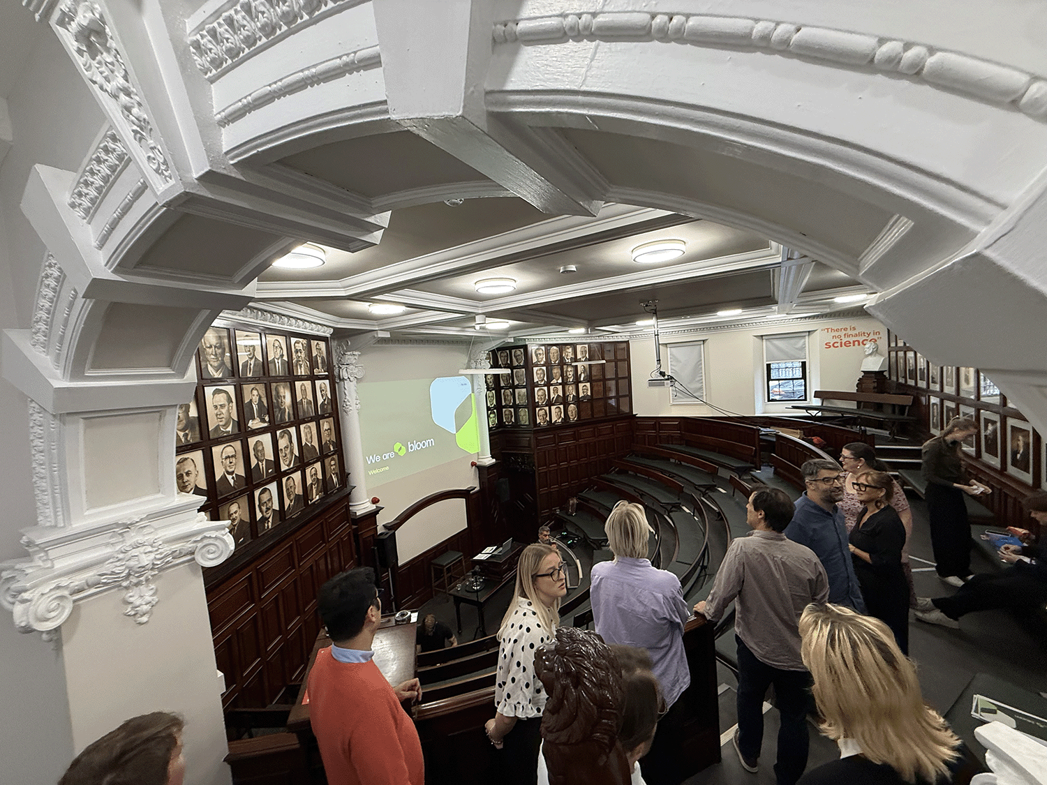 A 19th Century style lecture hall with white painted stone arches and wooden seating is being visited by a group of researchers who look around.