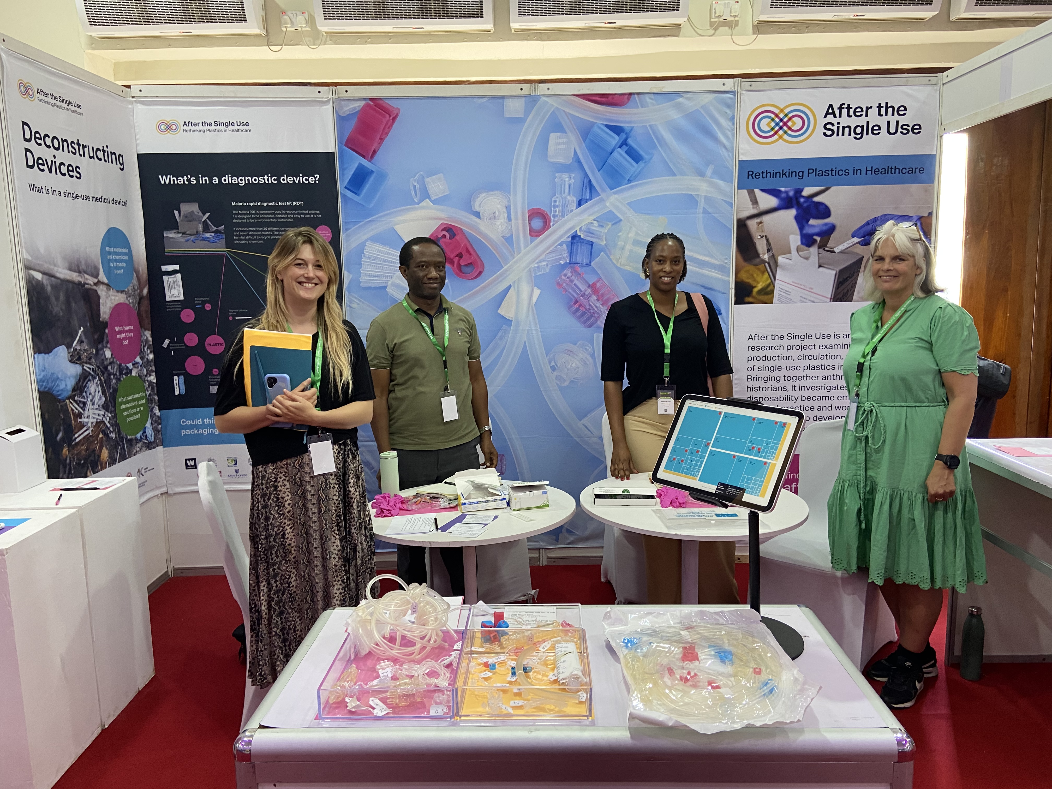Four researchers stand in an exhibition stand for After the Single Use. There's a table infront of them with perspex boxes of plastic medical equipment. 