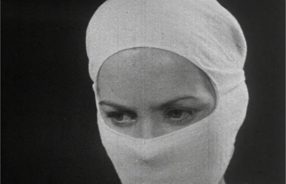 A black and white still from a medical film. The nurse pictured has a mask covering the face and hair, only the eyes of the person are shown in concentration.  