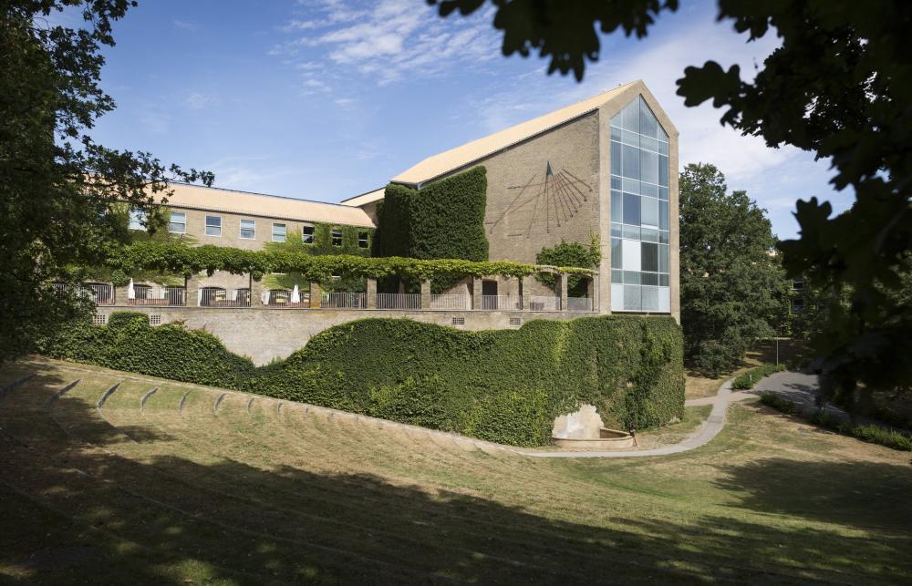 A large grey university building with a large glass facade and mid century brick details is photographed amongst some leafy trees and lush green bushes. 