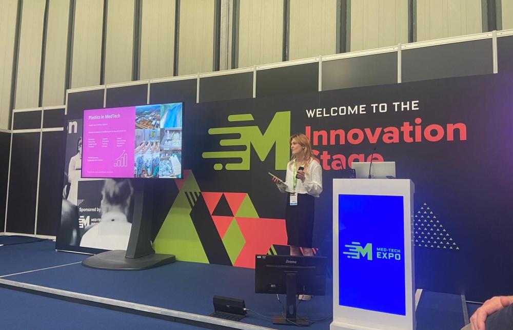 An exhibition presentation stage with screen and large dark coloured backdrop. In front is a researcher presenting with a microphone.  