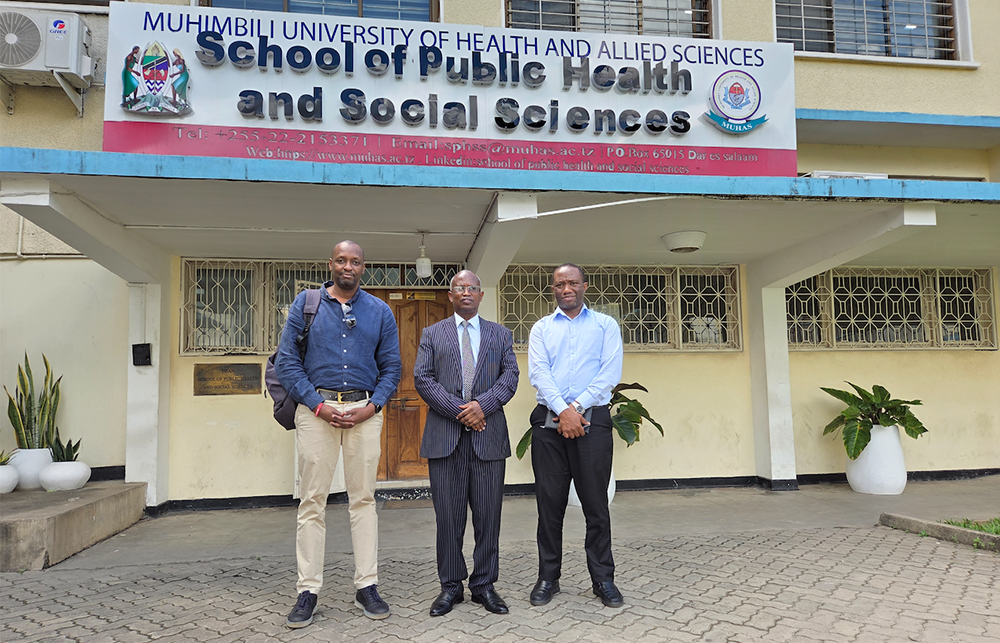Researchers and representatives stand outside the School of Public Health and Social Sciences in Muhimbli. The building has a marigold plaster exterior with decorative metal work on the windows. 