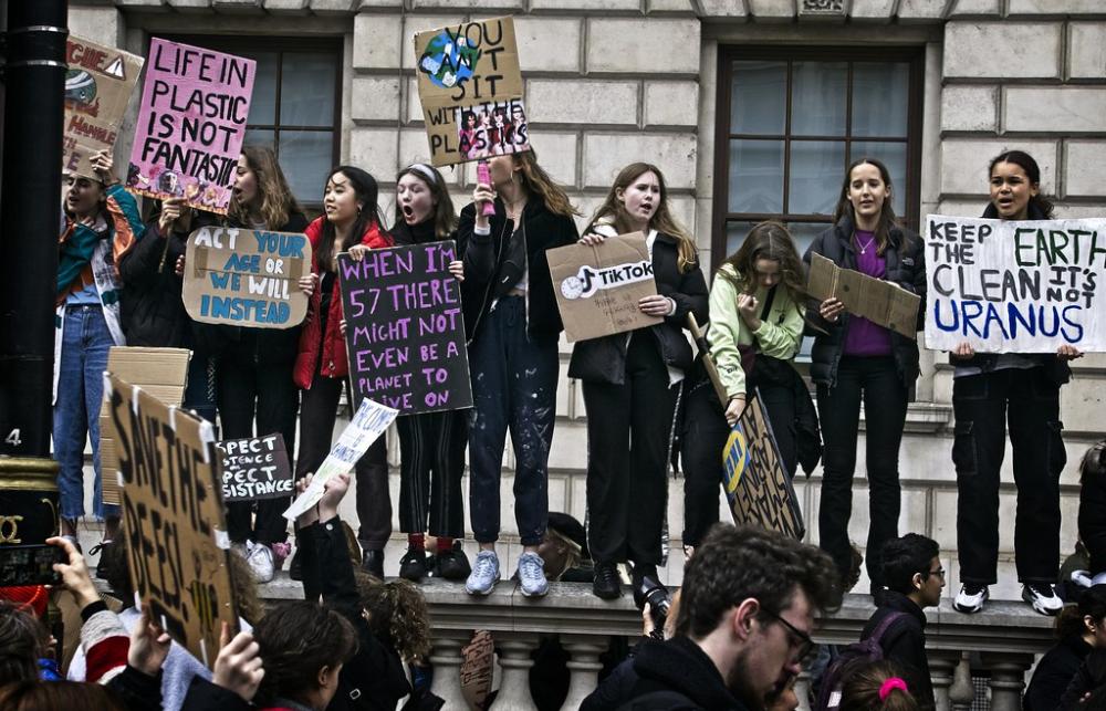 Young protesters stand on a stone wall with placards highlighting plastics and planetary health. 