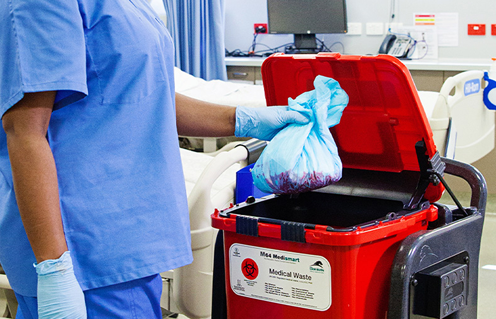 A hospital worker in blue scrubs puts a plastic bag of medical waste into a red bin. 