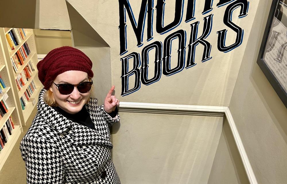 Miranda Melcher stands on a staircase pointing at the words "More Books" which have been printed on the wall by her side.